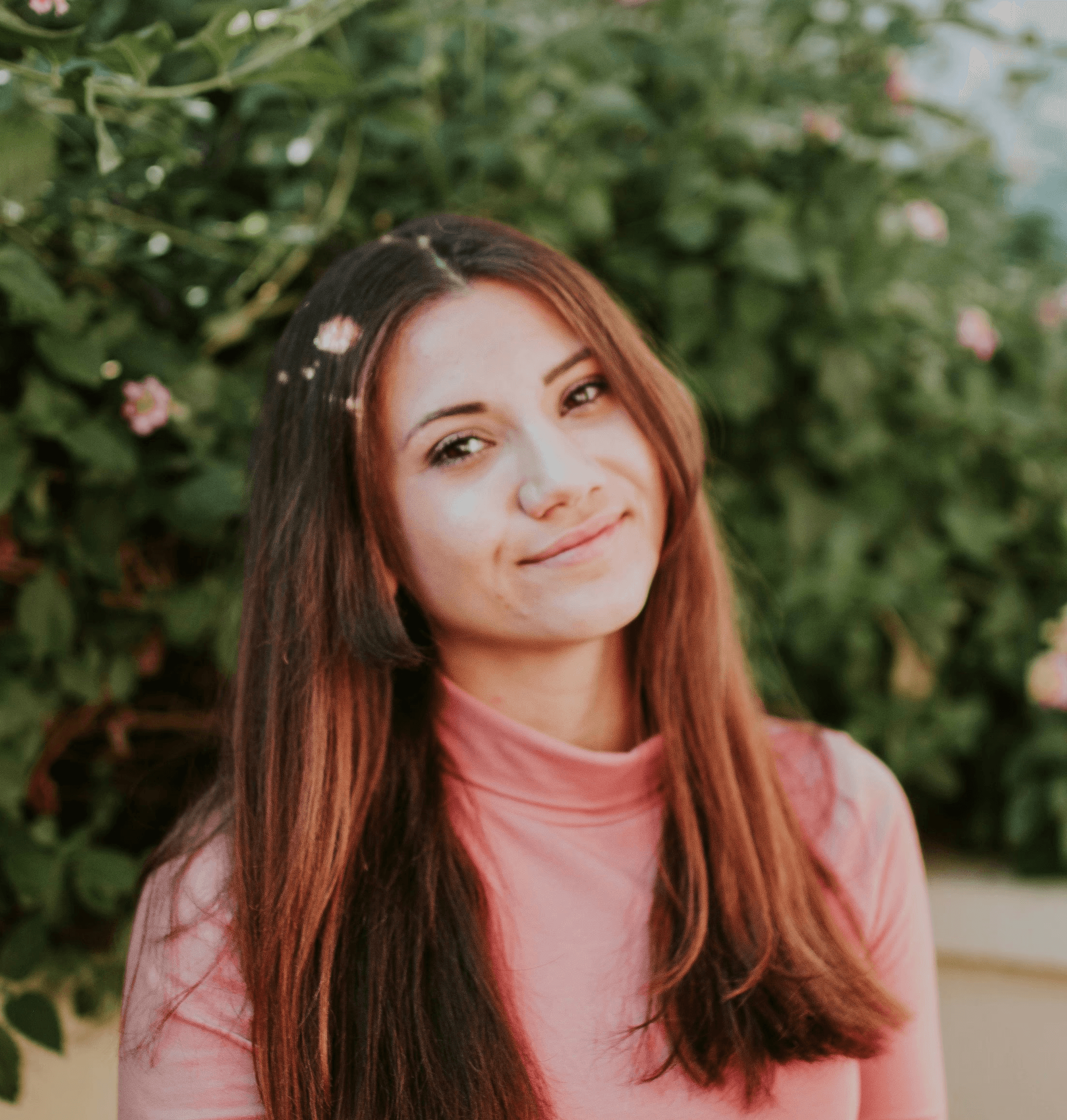 A young woman with long hair smiles softly, surrounded by greenery and delicate flowers in the background.